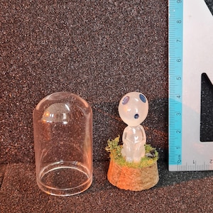 Puede incluir: Una campana de cristal transparente junto a una pequeña figura translúcida de un espíritu del bosque sentado, posada sobre una base de madera en miniatura con musgo verde. Una regla está en el fondo.