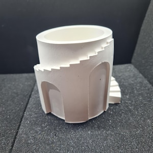 May include: A white, cylindrical planter with a unique architectural design. The planter features a spiral staircase and arched openings, creating a miniature building aesthetic. The interior is a smooth, open space for planting.