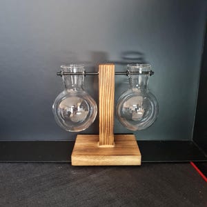 May include: A clear glass propagation station with two round bulbs, held by a wooden stand. The bulbs are suspended by black metal rods. The wooden base and central support have a warm, natural finish, creating a minimalist aesthetic.