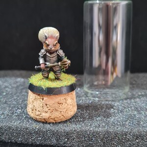 Peut inclure: Figurine miniature fantastique d'un écureuil guerrier, vêtu d'une armure et tenant une arme. La figurine est posée sur une base en liège avec de l'herbe artificielle, à côté d'un cylindre en verre transparent. L'écureuil a une fourrure brun clair.
