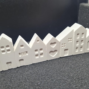 Può includere: Una fila di piccole case in ceramica bianca con diverse forme di tetto e motivi di finestre. Alcune case presentano ritagli a forma di cuore, altre finestre rettangolari. Le case sono disposte in una linea curva.