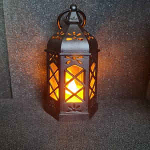 Pode incluir: Lanterna de metal preto com uma luz quente e brilhante no interior. A lanterna tem um padrão floral decorativo e uma alça. A luz brilha através de recortes geométricos, criando um ambiente acolhedor. É um item decorativo.