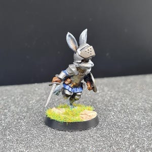 以下が含まれることがあります： 鎧をまとったウサギの騎士のミニチュアフィギュア。ヘルメット、胸当て、青いスカートが含まれています。ウサギは剣と盾を持ち、草と岩のある台座の上に立っています。フィギュアは詳細な陰影で塗装されています。