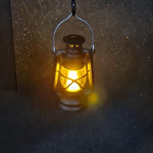 May include: A small, black lantern with a bright yellow light inside, hanging from a black chain. The lantern has a silver handle and a classic design. The background is dark and textured, with a warm glow around the lantern.