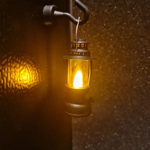 May include: A small, black lantern with a warm, yellow light hanging from a matching hook. The lantern has a classic design with a glass enclosure and a decorative top. The hook is mounted on a dark surface, creating a cozy, inviting ambiance.