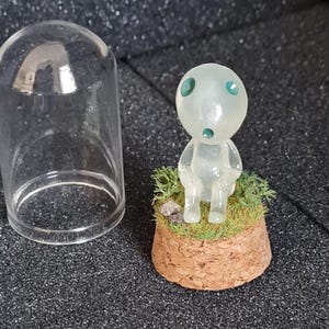 May include: A translucent figurine of a forest spirit with green eyes and mouth, sitting on a cork base with faux grass and a small rock. A clear glass dome sits to the left. The figurine is approximately 2 inches tall.