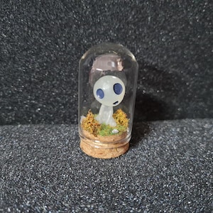 Puede incluir: Un terrario de vidrio en miniatura con una figura del espíritu Kodama. La figura tiene un cuerpo blanco y ojos azules, en medio de musgo preservado y follaje seco. El terrario tiene una base de corcho.