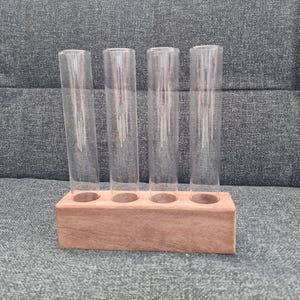 Könnte beinhalten: Ein Holzblock mit vier Löchern hält vier klare Reagenzgläser. Die Reagenzgläser sind zylindrisch und scheinen leer zu sein. Der Holzblock ist hellbraun.