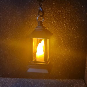 Puede incluir: Una pequeña linterna negra con una luz amarilla cálida en su interior, colgada de una cadena de metal. La linterna tiene una forma rectangular clásica con un techo puntiagudo. El fondo es oscuro y salpicado de oro.