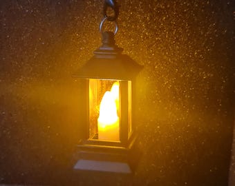 May include: A miniature black lantern with a warm, yellow glow emanating from within, suspended by a metal chain. The lantern features a traditional, rectangular design with a pitched roof. The background is dark and speckled with gold.