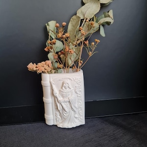 Puede incluir: Un jarrón blanco de cerámica con forma de libro con un relieve de la Parca sosteniendo una guadaña. El jarrón contiene flores secas y hojas de eucalipto. El jarrón está colocado contra una pared gris oscura.