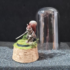 Puede incluir: Figura en miniatura de un guerrero ardilla, vestido con armadura y empuñando una espada y un escudo. La figura está colocada sobre una base de corcho con césped artificial y se exhibe bajo una campana de cristal. La ardilla tiene una cola marrón y blanca.
