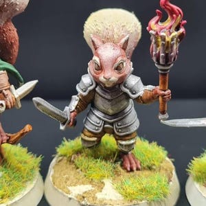 May include: A hand-painted miniature squirrel warrior figurine. The squirrel is clad in silver armour and holds a sword and a torch with a red and yellow flame. It stands on a small, round base with green and yellow grass. The figurine is highly detailed.
