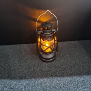 May include: A small, black lantern with a clear glass enclosure and a silver handle. The lantern is lit with a warm, orange glow, suggesting a candle or small light source inside. The lantern is a miniature version of a classic design.