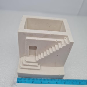 Peut inclure: Un petit pot de fleurs en béton blanc cassé avec un design architectural unique. Le pot a une forme de cube avec un détail d'escalier sur le côté. Le pot mesure environ 7.6 cm de large.