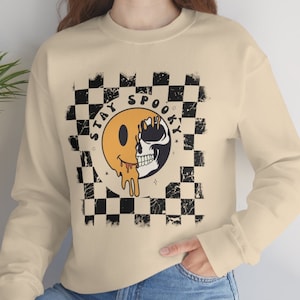 Könnte beinhalten: Ein beiger Pullover mit einem schwarz-weißen Karomuster und einer Grafik eines schmelzenden Smiley-Gesichts mit einem Schädel darin. Die Grafik ist von einem schwarzen Kreis mit dem Text "Stay Spooky" in Weiß umgeben.
