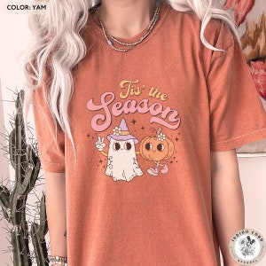 Könnte beinhalten: Ein orangefarbenes T-Shirt mit einem Aufdruck eines Geistes, der einen Hexenhut trägt, und einem Kürbis mit einer Blume auf seinem Stiel. Der Text "Tis' the Season" ist in einer Retro-Schrift über dem Aufdruck geschrieben.