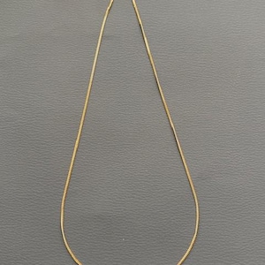 May include: A delicate gold-colored chain necklace. The chain is thin and appears to be made of small, rectangular links. The necklace is displayed against a gray background, highlighting its simple elegance.