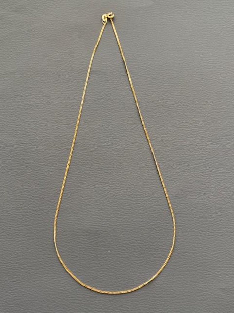 May include: A delicate gold-colored chain necklace. The chain is thin and appears to be made of small, rectangular links. The necklace is displayed against a gray background, highlighting its simple elegance.