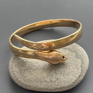 Pode incluir: Uma pulseira de metal em tom dourado com o formato de uma cobra enrolada. A superfície apresenta um padrão de diamante texturizado ao longo do corpo e da cabeça, com uma pequena pedra vermelha escura como olho. Acabamento liso e polido.