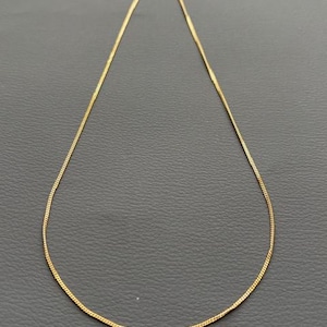 May include: A delicate gold-colored chain necklace. The necklace is thin and appears to be made of small, interconnected links. The chain is displayed against a dark gray background.
