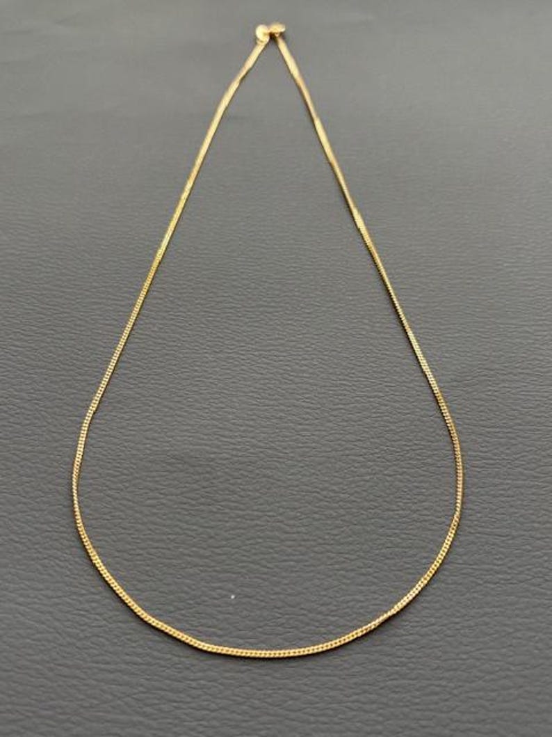 May include: A delicate gold-colored chain necklace. The necklace is thin and appears to be made of small, interconnected links. The chain is displayed against a dark gray background.
