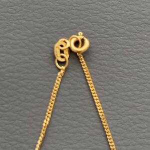 May include: Close-up of a gold-colored necklace chain with a lobster clasp. The chain is made of small, interconnected links, and the clasp is a simple, secure design. The necklace is displayed against a gray background.