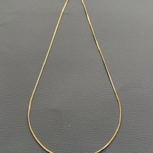 May include: A delicate gold-colored chain necklace. The necklace is thin and appears to be made of small, connected links. The chain is displayed against a dark gray background, highlighting its simple elegance. A classic jewelry piece.