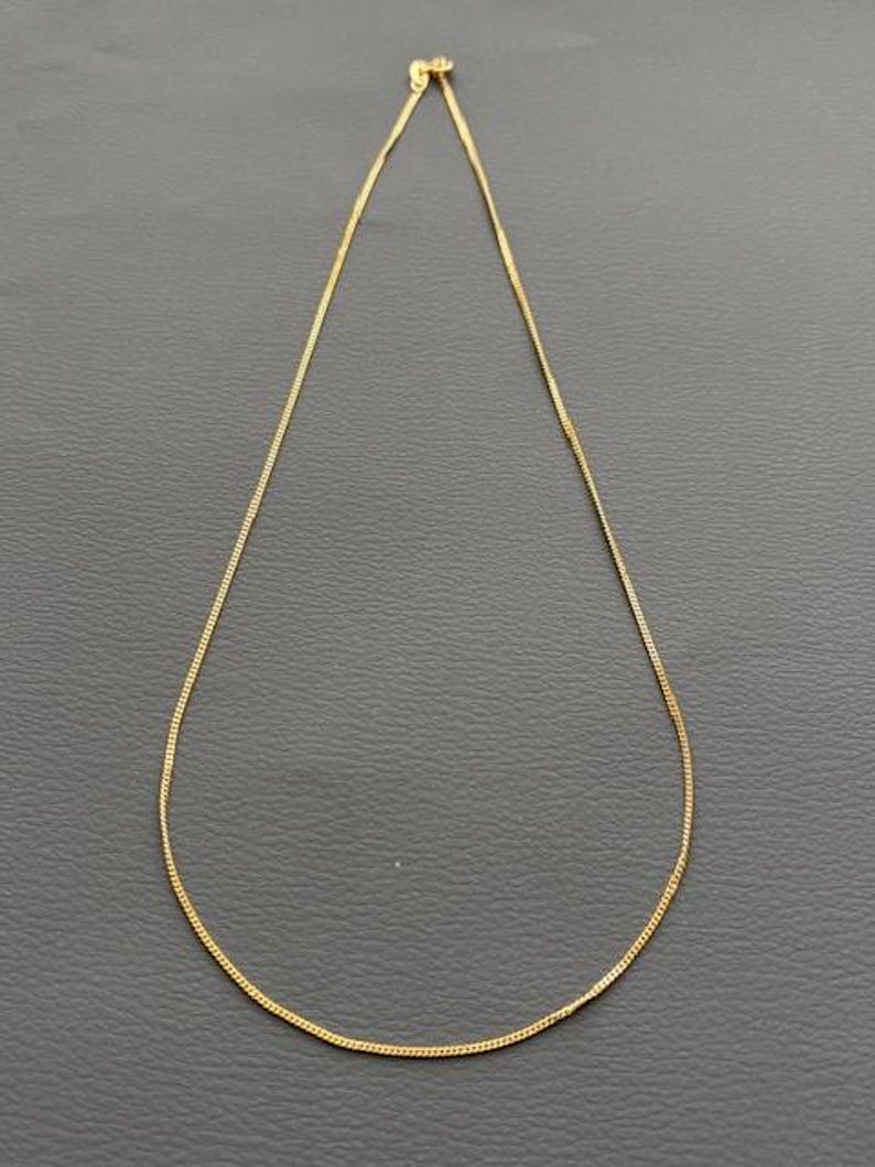 May include: A delicate gold-colored chain necklace. The necklace is thin and appears to be made of small, connected links. The chain is displayed against a dark gray background, highlighting its simple elegance. A classic jewelry piece.