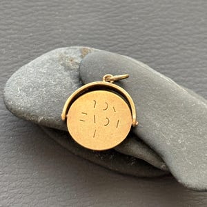 May include: A circular gold-toned metal pendant featuring a rotating centre disc held within a curved frame. The flat surface of the disc displays engraved markings that resemble stylised Hebrew characters or abstract symbols, with a small jump ring at the top.