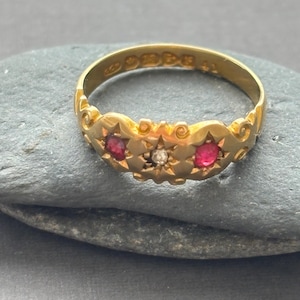 May include: A gold ring featuring a central clear stone flanked by two small red stones. The band has an ornate, scalloped edge design with engraved details and visible hallmarks stamped on the interior surface of the metal band.