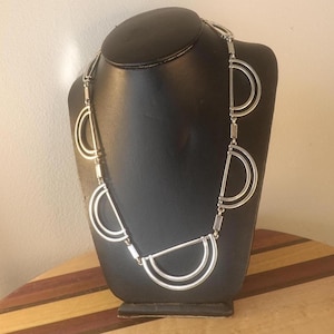 Könnte beinhalten: Eine silberfarbene Halskette mit geometrischem Design, die mehrere halbkreisförmige Formen aufweist, die durch rechteckige Glieder verbunden sind. Die Halskette wird auf einer schwarzen Schaufensterpuppe präsentiert.