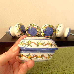May include: A collection of ceramic cabinet knobs and a small container. The knobs have a white base with blue and yellow floral designs. The container is white with a blue rim and a similar floral pattern. The knobs are attached to metal screws.