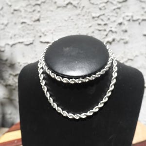 May include: A silver-colored rope chain necklace displayed on a black jewelry bust. The necklace is made of intertwined links, creating a textured, braided appearance. The bust is set against a textured, light-colored wall.