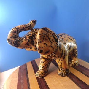 Könnte beinhalten: Eine Keramik-Elefantenfigur mit einem tan- und schwarz gefleckten Muster. Der Elefant hat seinen Rüssel erhoben und steht auf einer Holzoberfläche vor blauem Hintergrund. Die Figur ist ein dekoratives Objekt.