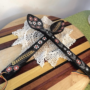 May include: Two black wooden spoons crossed over each other, featuring hand-painted floral motifs in red, white, and gold. The handles are decorated with wheat stalks and small flowers, and each end has a thin, multi-colored ribbon loop attached.