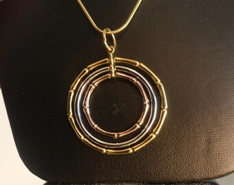 May include: A gold, silver, and rose gold necklace with a pendant featuring three concentric circles.