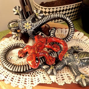 May include: Three ceramic lizard ornaments. One is red with black stripes, and the other two are black with white and yellow floral patterns. The lizards are displayed on a wooden surface with a white lace doily.