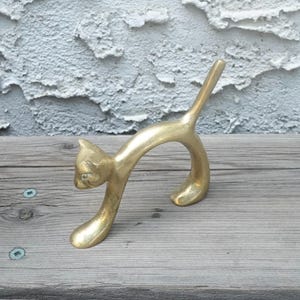 Könnte beinhalten: Eine goldfarbene Messingkatzenfigur in spielerischer Pose, mit gewölbtem Rücken und erhobenem Schwanz. Die Katze hat ein detailliertes Gesicht und steht auf einer verwitterten Holzoberfläche. Die Skulptur ist ein dekorativer Artikel.