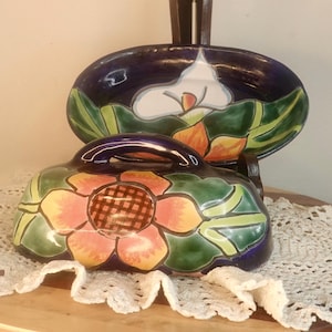 May include: A ceramic serving dish and lid set with a floral design. The set features a dark blue background with hand-painted flowers in orange, yellow, and green. The lid has a handle and the set is placed on a lace doily.