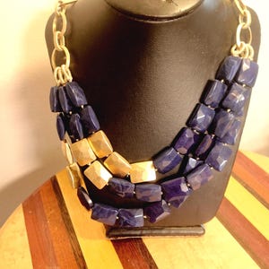 May include: A multi-strand necklace with gold-tone chain and faceted, geometric beads. The necklace features alternating sections of navy blue and gold-colored beads. The necklace is displayed on a black mannequin.