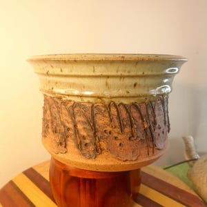 May include: A handcrafted ceramic planter with a light green upper section and a textured, brown lower section. The planter sits on a wooden stand. The design features a unique, organic pattern, perfect for displaying plants.