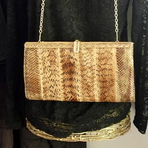 May include: A rectangular handbag with a snakeskin pattern in shades of brown and tan. The bag has a gold-coloured metal frame and a chain strap. The clasp is a small, silver-coloured rectangle. The bag is a vintage style.