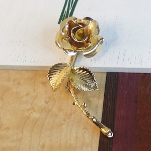 May include: A gold-coloured rose brooch with detailed petals and leaves. The stem and leaves have intricate vein detailing. A classic floral accessory.