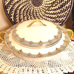 Op de afbeelding: Een crèmekleurige keramische serveerschaal met bijpassend deksel, voorzien van een decoratieve rand met lichtblauwe, gouden en roze bloemaccenten. De schaal heeft twee goudkleurige handgrepen aan de zijkanten en een goudkleurige knop op het deksel.