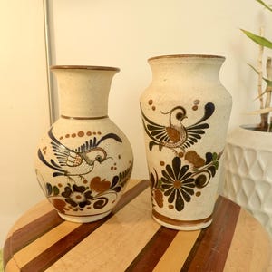 Op de afbeelding: Twee keramische vazen met handgeschilderde bloemen- en vogelmotieven. De hogere vaas heeft een bruin, blauw en wit ontwerp met een vogel en bloemen. De lagere vaas heeft een bruin, blauw en wit ontwerp met een vogel en bloemen.