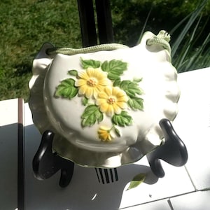 Può includere: Una tasca da parete in ceramica bianca con bordo smerlato, con margherite gialle in rilievo e foglie verdi. Un cordoncino verde è attaccato per appenderla. La tasca è esposta su un supporto nero.