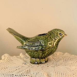Może przedstawiać: Ceramiczny wazon w kształcie ptaka w odcieniach zieleni. Ptak ma szczegółowe tekstury piór i mały otwór na grzbiecie. Wazon stoi na małej, dekoracyjnej podstawie. Dziób ptaka jest jasnożółty.