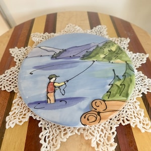 Puede incluir: Un plato de cerámica pintado a mano con una escena de paisaje azul y verde. Una persona de rojo está pescando en un lago con montañas en el fondo. El plato tiene un borde marrón y está decorado con un patrón de troncos.
