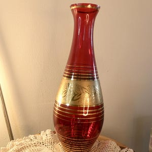 Pode incluir: Um vaso alto de vidro vermelho com detalhes dourados. O vaso apresenta listras douradas horizontais e uma faixa dourada decorativa com um desenho de folhas. A borda e a base também são adornadas com ouro.